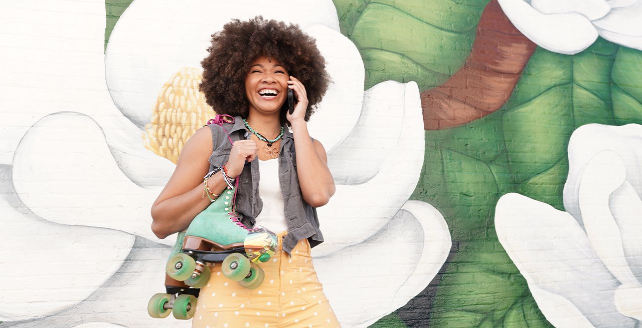 Woman holding roller skates and using her mobile phone, smiling, with colorful mural background